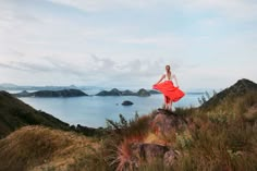 a woman in a red dress standing on top of a hill next to the ocean