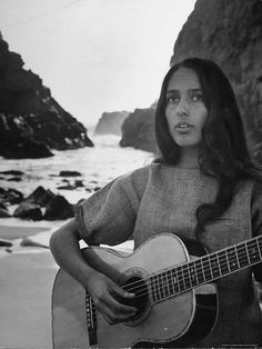 a young woman holding an acoustic guitar on the beach in front of rocks and water