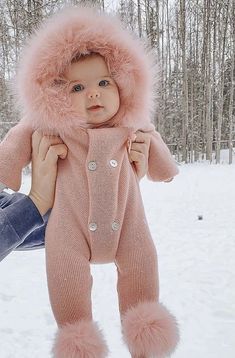 a person holding a baby wearing a pink snowsuit in the snow with trees in the background