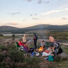 a group of people sitting around a campfire on top of a hill with mountains in the background