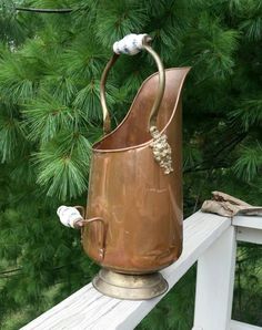 an old copper pitcher sitting on top of a white wooden porch next to pine trees