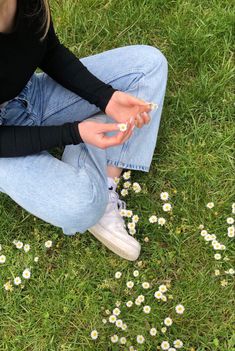 a woman sitting in the grass with daisies on her feet and one hand holding something