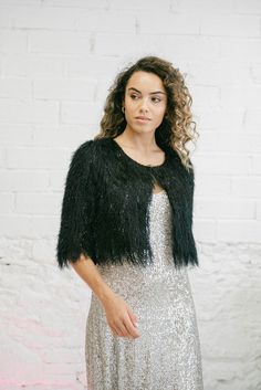 a woman standing in front of a brick wall wearing a silver dress and black jacket