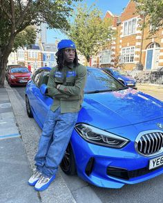 a man standing next to a blue car on the side of the road with his arms crossed