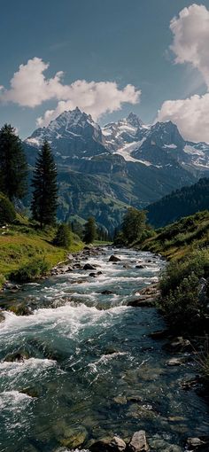 a river running through a lush green hillside covered in snow capped mountain range behind it