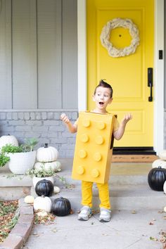 a young boy dressed up as a lego man outside his house for halloween with yellow door and black pumpkins