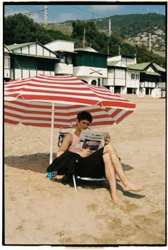 a man sitting under an umbrella on the beach reading a book while holding a magazine