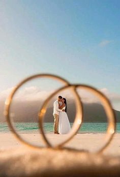 a bride and groom standing on the beach with their wedding rings in front of them