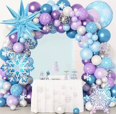 a table topped with balloons and snowflakes next to a large arch covered in blue, white and purple decorations