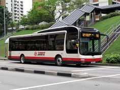 a red and white bus driving down a street next to a lush green hillside covered in trees