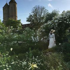 a woman wearing a white dress and hat in a garden with tall buildings behind her