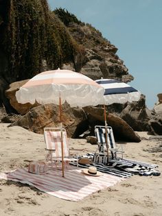 two lawn chairs and an umbrella on the sand at the beach with rocks in the background