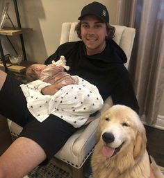 a man sitting in a chair next to a dog and holding a baby on his lap