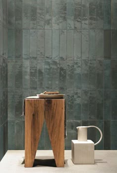 a wooden table sitting next to a white vase on top of a counter in front of a green tiled wall