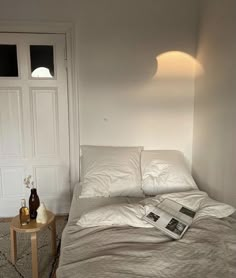 a bed with white sheets and pillows next to a wooden side table in front of a door