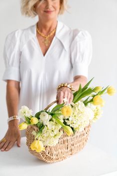 a woman is holding a basket with flowers in it on a white table and smiling