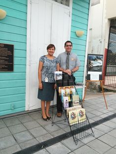 two people standing in front of a blue building with signs on it and pictures behind them