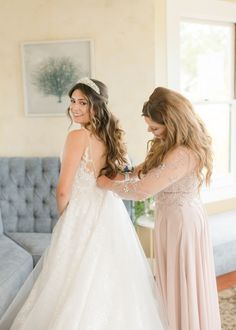 two women standing next to each other in front of a couch and one is wearing a wedding dress