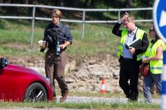 two people standing next to a red car on the side of a road with traffic cones around them