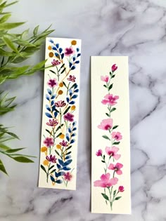 two bookmarks with watercolor flowers and leaves on them next to a potted plant