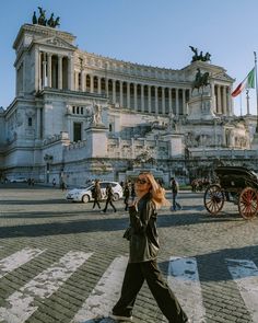 a woman walking across a cross walk in front of a building with statues on it