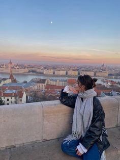 a woman sitting on top of a cement wall next to a cityscape at sunset