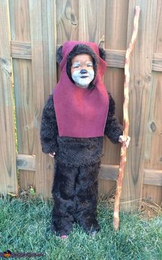 a child dressed as a bear holding a stick and wearing a costume that looks like it is standing in front of a fence