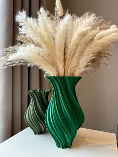 two green vases with white feathers in them on a table next to a window