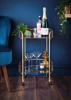 a gold bar cart with two glasses and bottles on it next to a blue chair