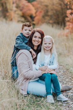 a mother and her two children sitting on the ground in an open field with fall foliage