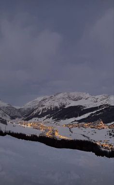 snow covered mountains with buildings and lights in the distance under a cloudy sky at night