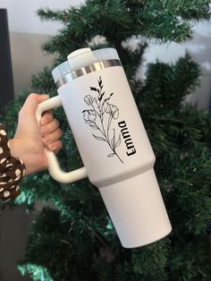 a person holding a white coffee cup in front of a christmas tree with flowers on it