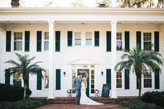 a bride and groom standing in front of a large white house with green shutters