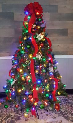 a decorated christmas tree with red ribbon and lights on it in front of a wooden wall