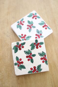 two ceramic coasters with holly and red berries on them sitting on a wooden table