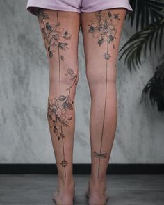 a woman's legs with flowers and vines tattooed on her leg, standing in front of a wall