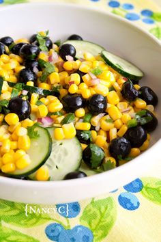 a white bowl filled with corn, cucumber and black olives on top of a table