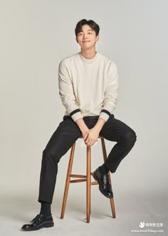 a man sitting on top of a wooden stool wearing a white sweater and black pants