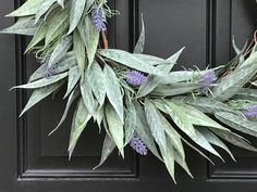 a wreath with purple flowers and green leaves hanging on a black front door, next to a pair of scissors