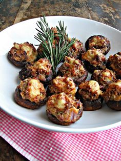 a white plate topped with stuffed mushrooms covered in cheese and seasoning next to a sprig of rosemary