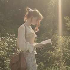 a woman standing in the grass holding a book and looking at it with sunlight shining on her face