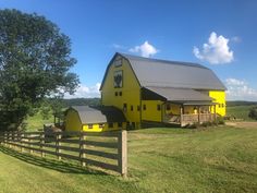 a large yellow barn sitting on the side of a lush green field next to a wooden fence