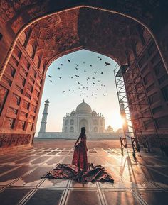 a woman is sitting on the ground in front of an archway with birds flying overhead