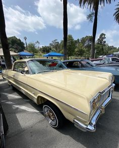 an old yellow car parked in a parking lot next to palm trees and blue umbrellas