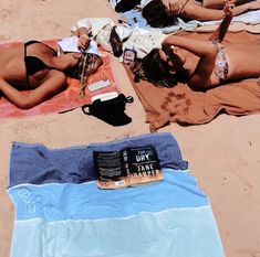 two women laying on towels in the sand next to each other and reading a book