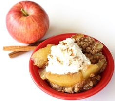 an apple and cinnamon crumbler in a red bowl next to an apple on a white table