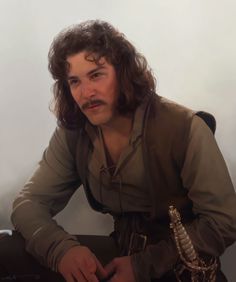 a man with long hair sitting down wearing a brown shirt and holding a knife in his hand