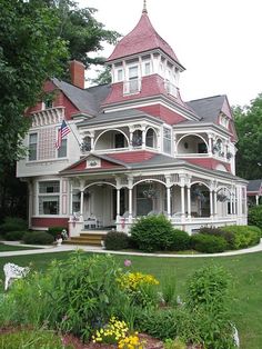 an old victorian style house with red roof and white trim on the front porch, surrounded by lush green trees