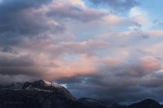 the mountains are covered in snow and clouds as the sun is setting behind them on a cloudy day