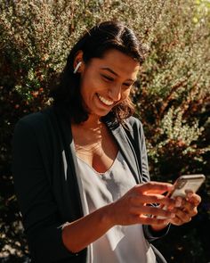 a woman smiles as she looks at her cell phone while standing in front of some bushes